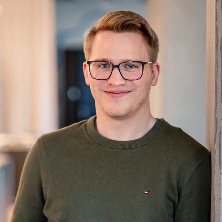 MERA-Mitarbeiter mit kurzem rotblondem Haar und Brille steht lächelnd im Hamburger Büro für Landschaftsarchitektur. Er trägt einen dunkelgrünen Pullover und lehnt seitlich an einer Wand; der Hintergrund ist hell und ruhig unscharf.