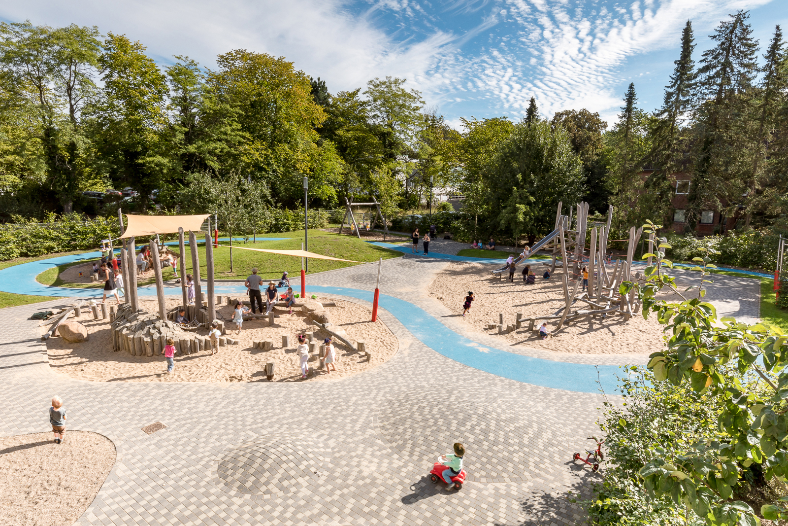 Perspektivische Sicht auf einen Großteil des Freigeländes der Kita Mose. Man sieht zwei Sand-Kletterspielbereiche, einen gepflasterten Vorplatz mit integrierten Spielhügeln und natürlich wirkende Holz-Klettergerüste in den Sandsielinseln. Dahinter weitläufige. hügelige Rasenfläche, alten Baumbestand und den blauen Gummigranulatweg, der sich durchs Gelände schlängelt.