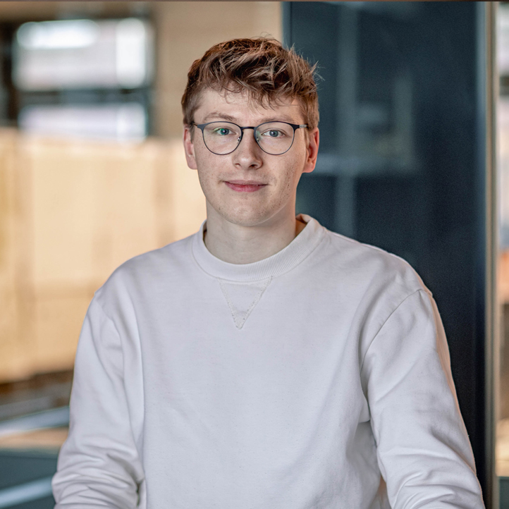 Junger MERA-Mitarbeiter mit kurzem rotblondem Haar und Brille sitzt im Hamburger Büro für Landschaftsarchitektur vor einem ruhig unscharfen Hintergrund. Er trägt ein helles Sweatshirt und blickt mit neutral-freundlichem Ausdruck direkt in die Kamera.