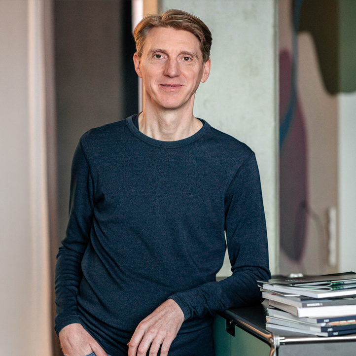 MERA-Mitarbeiter mit kurzem dunkelblondem Haar steht im Hamburger Büro für Landschaftsarchitektur an einem Tisch oder Tresen. Er trägt ein dunkelblaues Langarmshirt, blickt ruhig und freundlich in die Kamera; neben ihm liegt ein Stapel Unterlagen oder Zeitschriften, der Hintergrund ist hell und leicht unscharf.