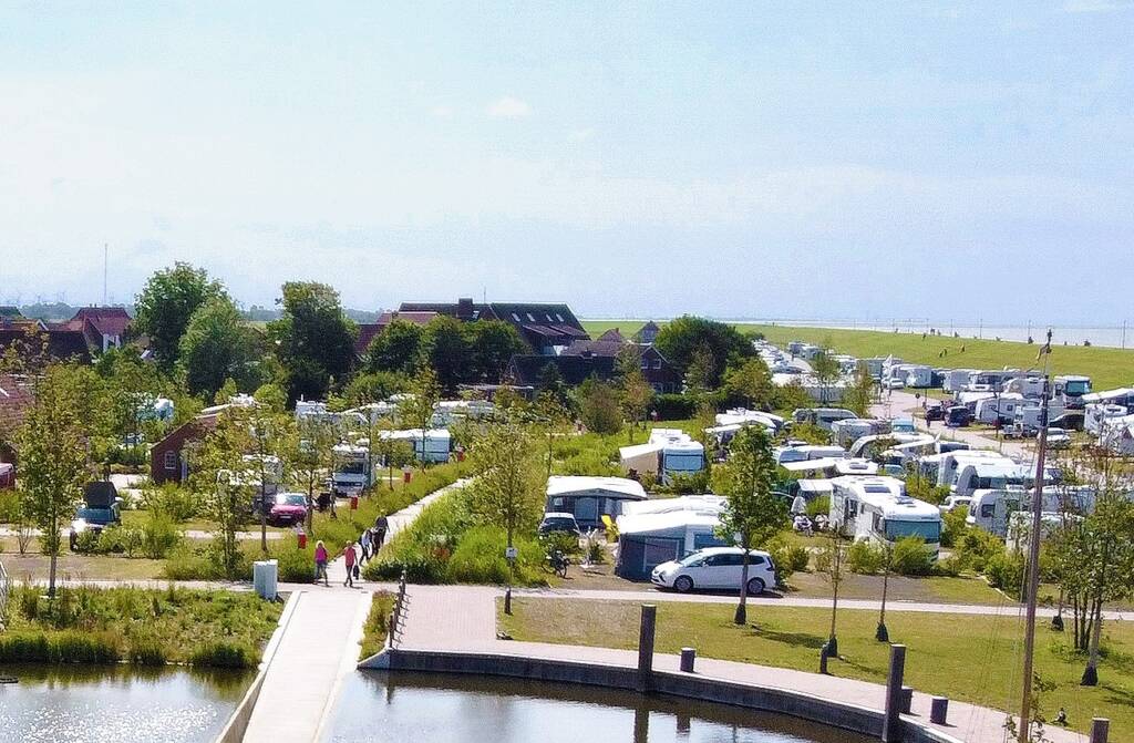 Auf dem Bild ist der Holzbohlenweg über den Campingplatz Neuharlingersiel aus der Luft aus Richtung Kurparksee zu sehen. Deutlich zu erkennen, der dichte Schilfbewuchs links und rechts des Weges und die danebenliegenden Stellplatzparzellen mit üppiger umgebender Begrünung. Die Sonne scheint, der Himmel spiegelt sich auf dem Kurparksee und die Stimmung ist sommerlich frisch.