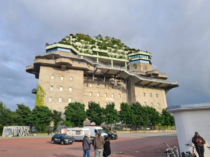 Zu sehen ist der ehemalige Flakturm an der Hamburger Feldstraße. Das Betonungetüm hat fünf Stockwerke obenauf erhalten, die üppig begrünt sind. Diese sind als grün bewachsene Terrasse von unten zu sehen. Im Vordergrund vor dem bunker eine Baumgruppe, davor roter Asphalt, darauf parken ein PKW und ein Caravan. Im Vordergrund sind einige Personen zu sehen.