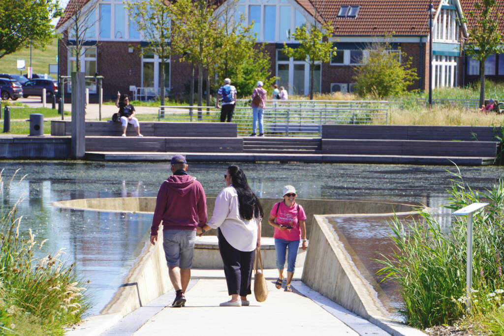 Das Foto zeigt das Seebauwerk im Kurpark Neuharlingersiel, ein besonderes Element der von MERA gestalteten Anlage. Ein geschwungener, abgesenkter Weg aus hellem Beton führt direkt in den See hinein und endet in einer runden Plattform, die unterhalb des Wasserspiegels liegt. Besucher*innen laufen dort entlang, bleiben stehen und betrachten die Wasserfläche aus nächster Nähe. Im Bild sind mehrere Menschen zu sehen: vorne eine Frau im pinkfarbenen Shirt, hinter ihr ein Paar, das Richtung Plattform geht. Im Hintergrund befinden sich breite Holzstufen am Ufer, die als Sitz- und Aufenthaltsbereich dienen, sowie Bäume, Gräser und Gebäude mit roten Ziegeldächern. Die Gestaltung ermöglicht ein unmittelbares Naturerlebnis: Menschen können fast auf Augenhöhe mit der Wasseroberfläche stehen und Enten sowie Spiegelungen beobachten.