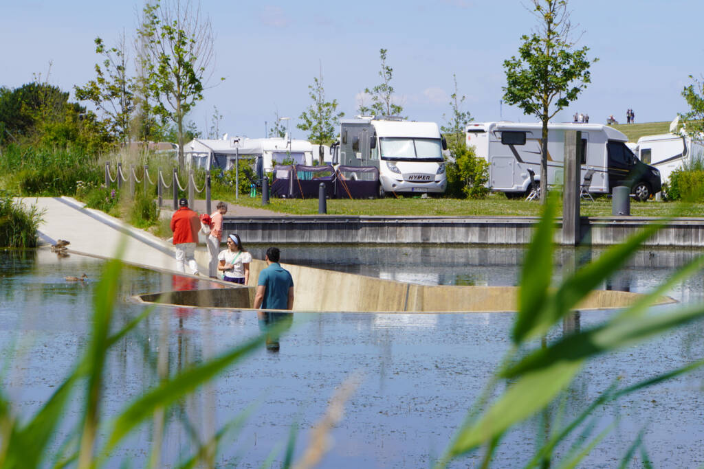 Das Foto zeigt den Kurparksee im Kurpark Neuharlingersiel, der von MERA neu gestaltet wurde. Im Vordergrund ist ein Wasserbecken zu sehen, durch das ein geschwungener Steg aus hellem Beton führt. Mehrere Menschen stehen darauf, unterhalten sich oder schauen auf die Wasserfläche, auf der auch Enten schwimmen. Im Hintergrund erkennt man Wohnmobile und Zelte des angrenzenden Campingplatzes, eingerahmt von neu gepflanzten Bäumen und Grünflächen. Die Gestaltung schafft eine Verbindung von Natur, Wasser und Freizeitnutzung – ein Ort, an dem Besucher*innen unmittelbar am Wasser verweilen können.