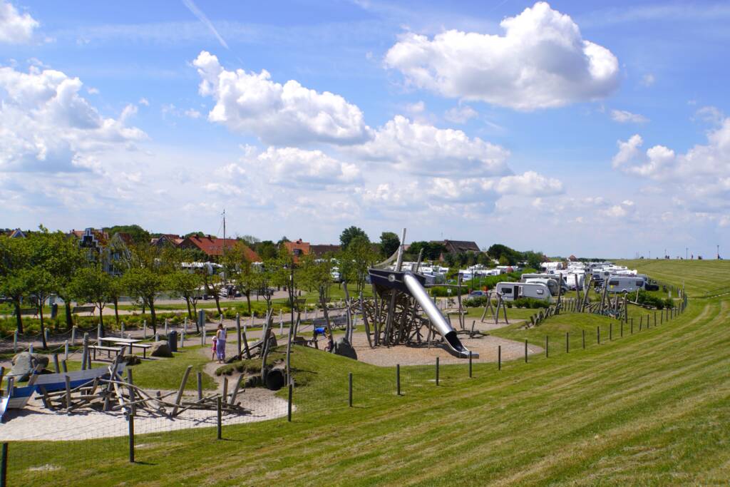 Das Foto zeigt den neuen Spielplatz im Kurpark Neuharlingersiel, der von MERA gestaltet wurde. Im Vordergrund zieht sich eine großzügige Spiellandschaft entlang des Deiches. Zu sehen sind verschiedene Spielgeräte aus naturbelassenem Holz – darunter Balancierstämme, Kletterelemente und ein großes Kletterschiff mit einer langen Edelstahlrutsche. Kinder spielen zwischen den Geräten, Erwachsene stehen daneben oder laufen vorbei. Im Hintergrund sind Bäume, Sitzbereiche und die angrenzenden Wohn- und Ferienhäuser erkennbar. Dahinter schließt sich der Campingplatz mit zahlreichen Wohnmobilen an, während sich rechts im Bild der grüne Deich hochzieht. Über allem spannt sich ein sommerlicher Himmel mit weißen Wolken. Die Szene vermittelt ein lebendiges Bild: Spiel, Aufenthalt und Landschaft greifen harmonisch ineinander – typisch für die landschaftsarchitektonische Gestaltung von MERA in Neuharlingersiel.