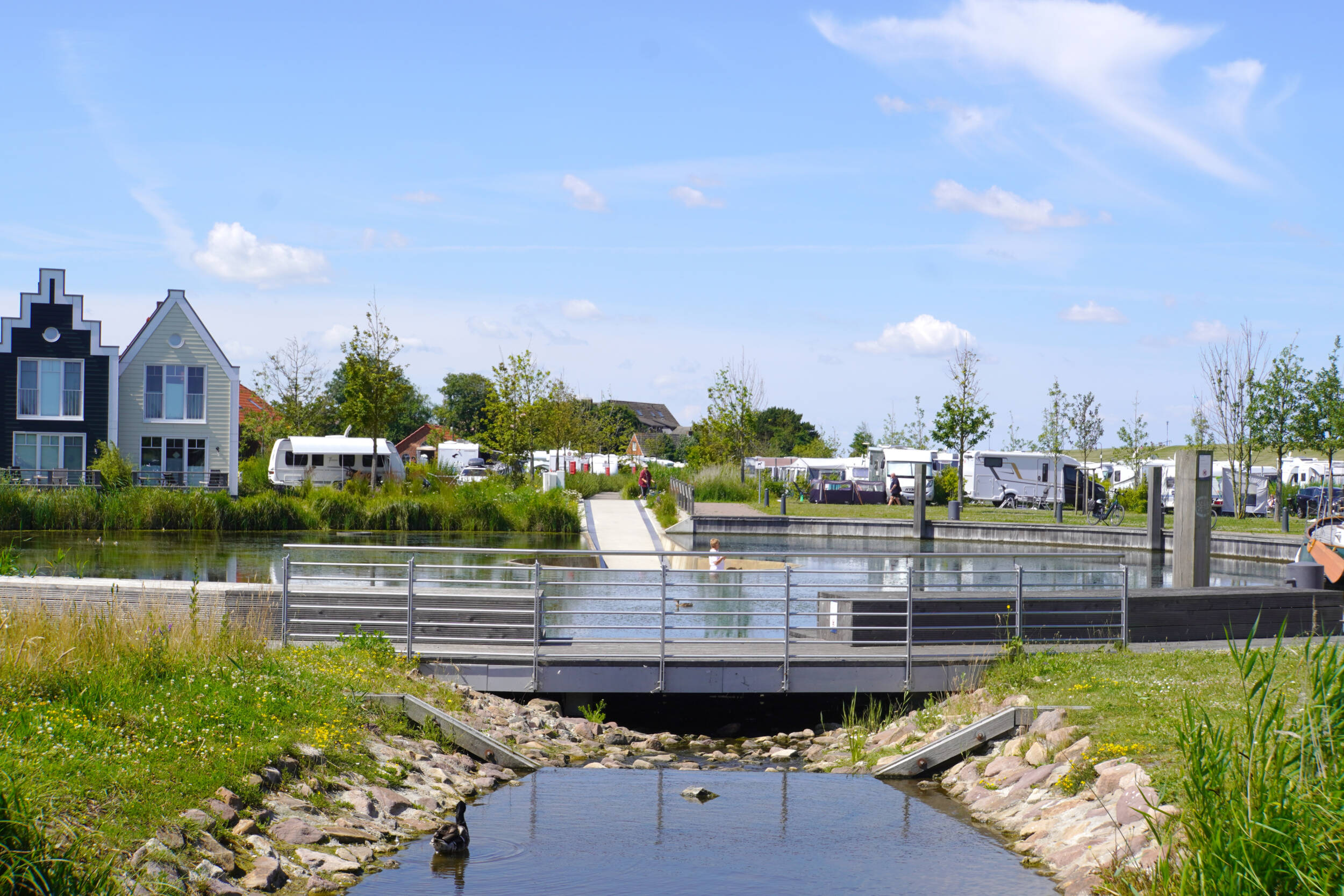 Fotografie einer neu gestalteten Parkanlage in Neuharlingersiel. Im Vordergrund ist ein Steingesäumter Wasserlauf zu sehen, rechts und links ist der Ufersaum Grasbewachsen. Quer durchs Bild eine Spaziergängerbrücke mit modernem Edelstahlgeländer. Dahinter sind Sitzpodeste rund um den dahinterliegenden Kurparksee zu erkennen. Im See, in der eingelassenen Seeplattform ist eine Person zu erkennen, die über die Wasseroberfläche schaut. Im Hintergrund die Rampe des des Seebauwerks, die zum Panoramasteg durch den Campingplatz führt. Im Hintergrund sind baumgesäumte Campingplätze mit Wohnwagen und Wohnmobilen zu erkennen. Das Wetter ist freundlich, der Himmel blau.