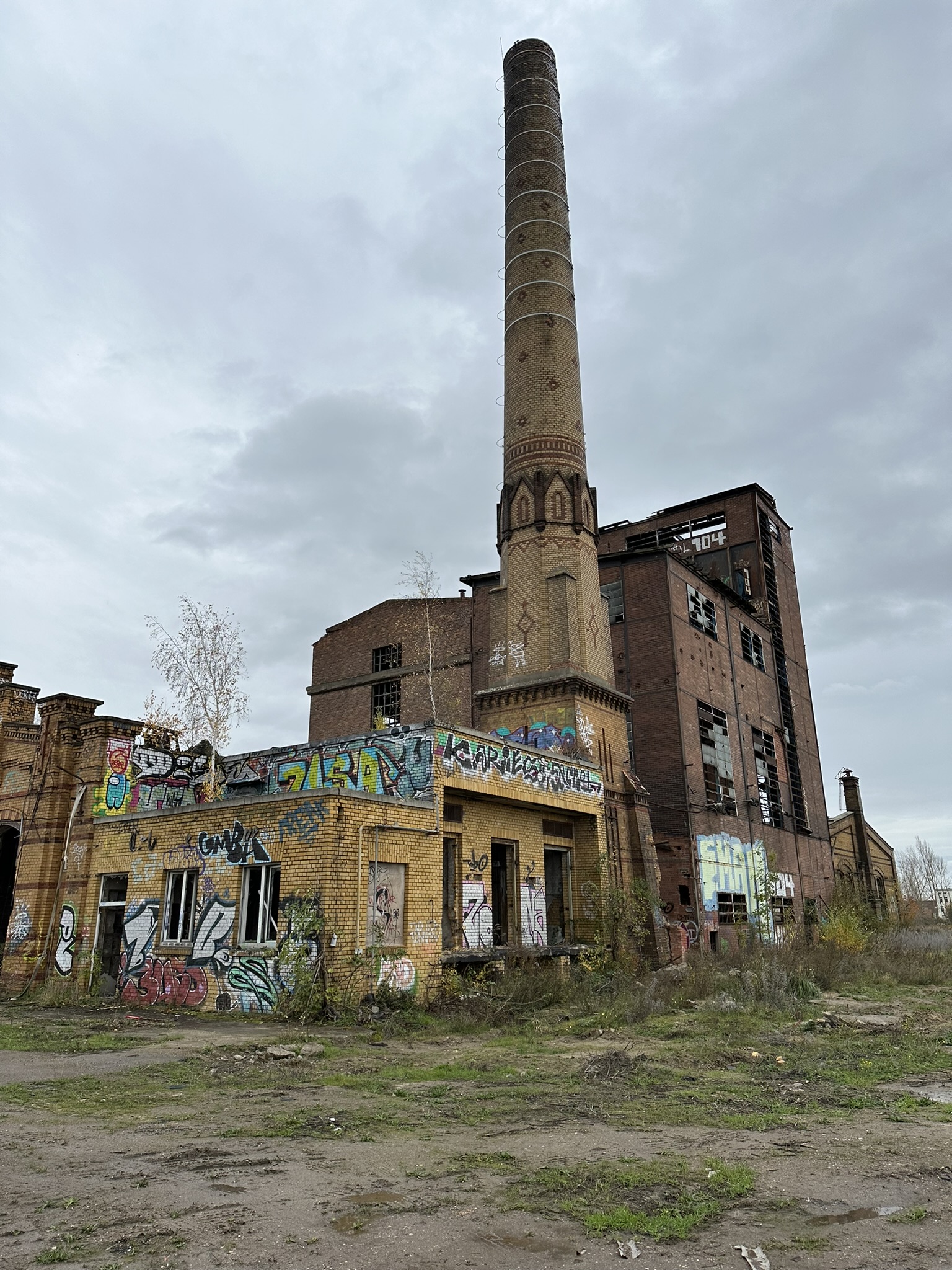 Altbestandsgebäude auf ehemaligem Eisenbahngelände. Zu sehen ist ein gemauerter Schornstein bzw. Turm sowie angrenzende Gebäude aus Gelbklinker, die mit Grafritti besprüht sind. Im Hintergrund ein mehrstöckiges Lagerhaus in Rotklinker. Davor zu sehen grasbewachsenes Industriebrachgelände.