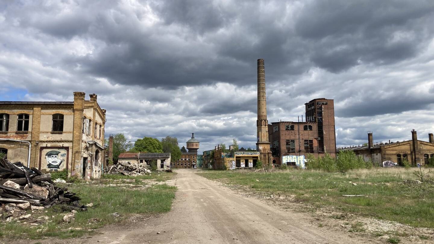 Im Mittelpunkt des Bildes verläuft ein breiter, unbefestigter Weg aus Erde und Schotter, der tief in das Gelände hineinführt. Links und rechts stehen stark gealterte Backsteingebäude, teils mit zerstörten Fenstern, offenen Fassaden und sichtbaren Spuren von Verfall. Auf der linken Seite liegen Trümmer und Bauschutt, dazwischen wächst vereinzelt Gras und niedrige Vegetation. Rechts ragen mehrere markante Industriebauten auf, darunter ein hoher, schmaler Schornstein, der das Areal weithin sichtbar prägt. Weitere Gebäude sind mit Graffiti versehen, einige wirken verlassen, andere nur teilweise erhalten. Im Hintergrund ist ein runder Wasserturm zu erkennen, der die industrielle Geschichte des Ortes unterstreicht. Über dem gesamten Areal liegt ein dramatischer Himmel mit schweren, dunklen Wolken, der die raue, offene Atmosphäre verstärkt. Das Bild vermittelt den Ausgangszustand eines großen Industriegeländes – geprägt von Weite, Geschichte und Potenzial für eine zukünftige landschaftsarchitektonische Entwicklung durch MERA.