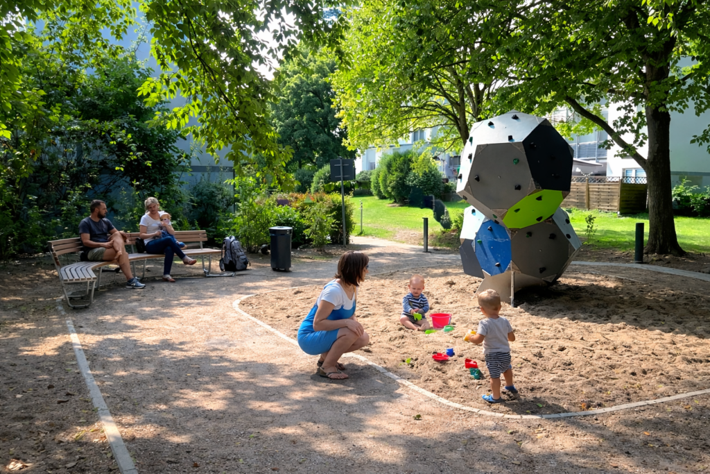 Ein sonniger Spielbereich in einer wohnungsnahen Grünanlage. In einem Sandspielbereich spielen zwei Kleinkinder mit bunten Sandspielzeugen, während eine Frau neben ihnen hockt und ihnen zuschaut. Hinter dem Sandbereich steht eine große geometrische Kletterstruktur aus mehrfarbigen Platten. Links sitzt eine Familie auf einer Bank unter Bäumen und beobachtet das Geschehen. Wege, Rasenflächen und Sträucher umgeben den Spielplatz, dahinter stehen mehrgeschossige Wohngebäude mit Balkonen.