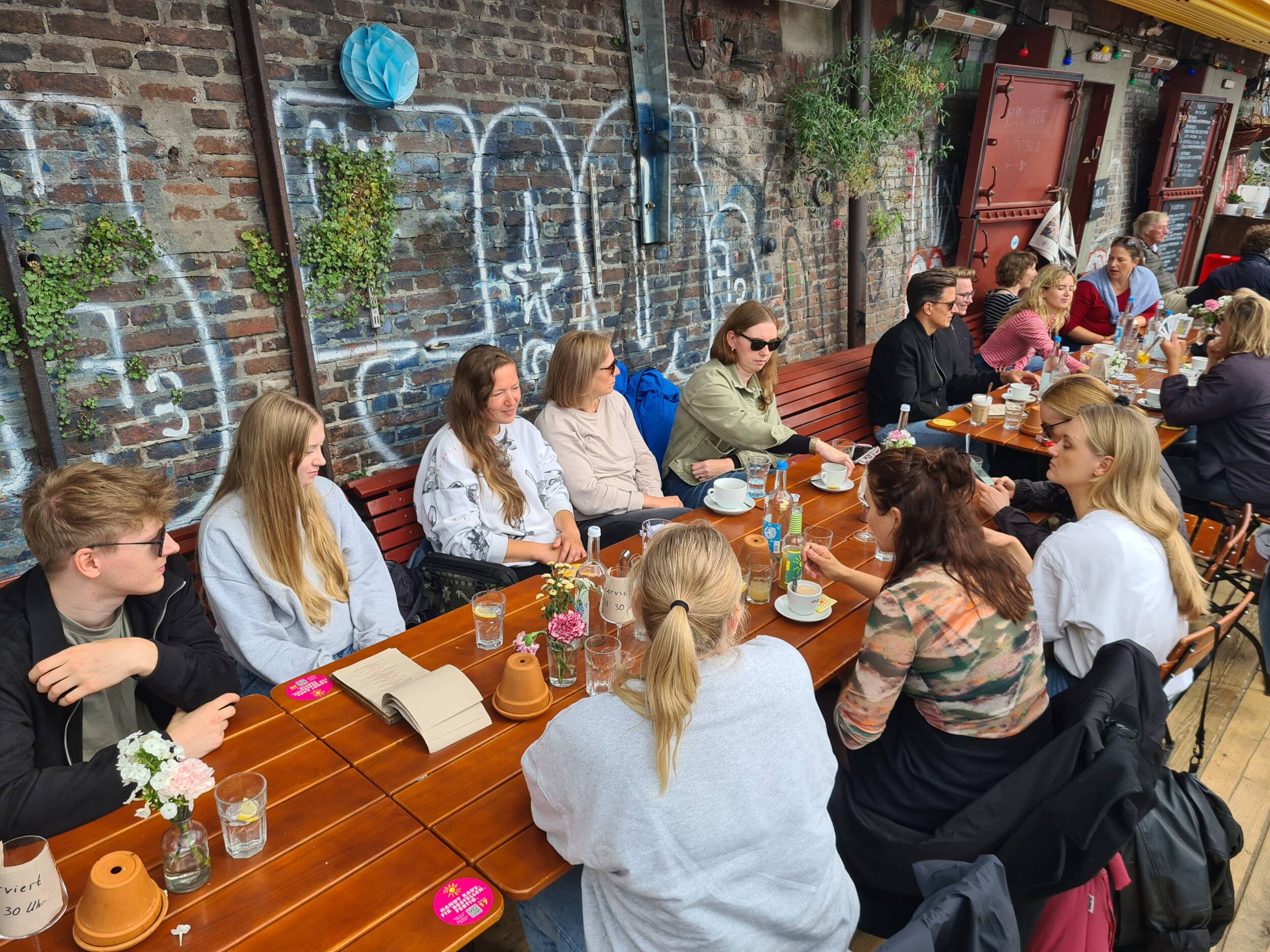 MERA-Mitarbeitende sitzen gemeinsam an einem langen Tisch im Außenbereich und tauschen sich in entspannter Runde aus. Die Gruppe ist vor einer bewachsenen Backsteinwand mit Graffiti zu sehen, auf dem Tisch stehen Gläser, Getränke und kleine Gegenstände; die Situation wirkt ungezwungen, lebendig und gemeinschaftlich.