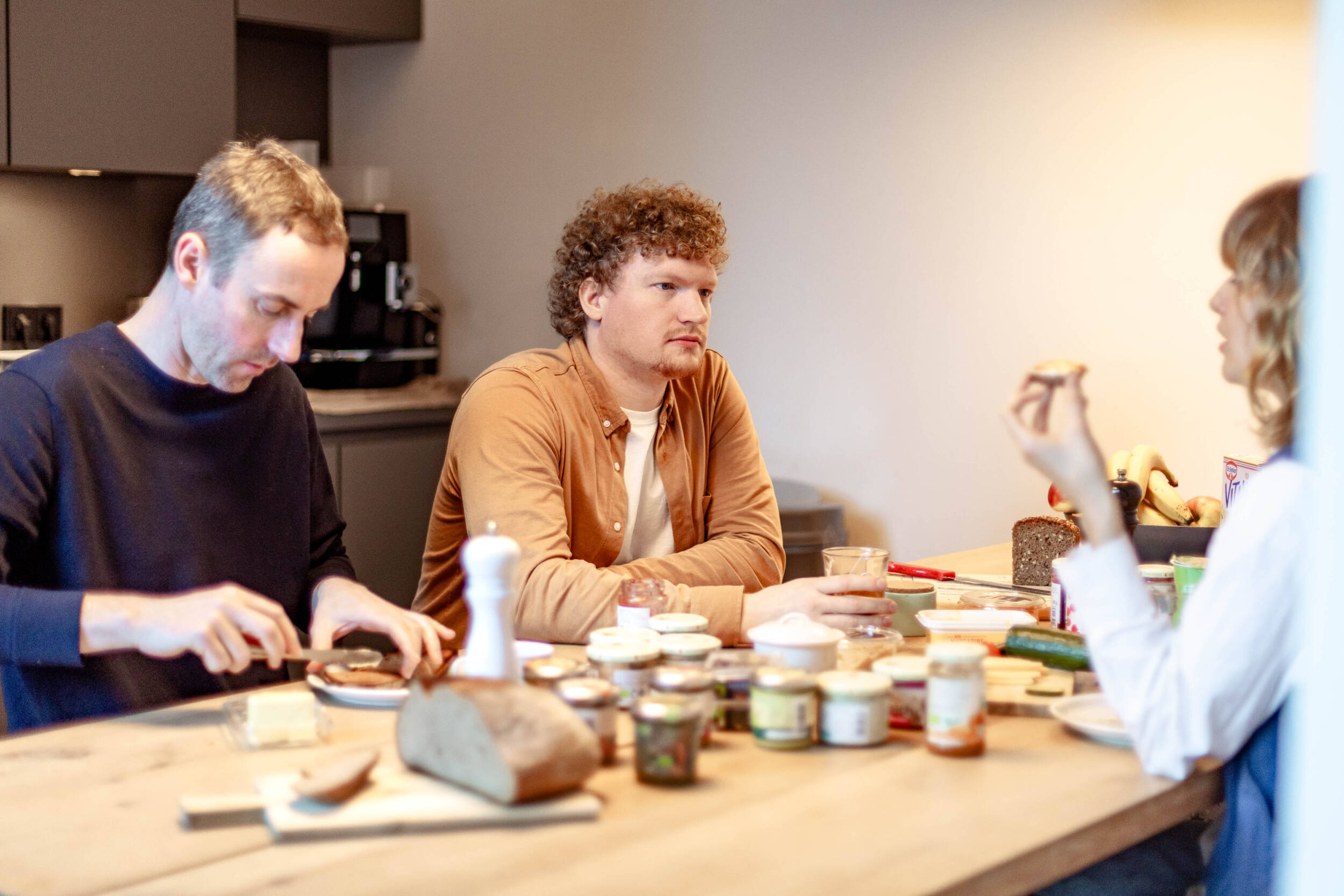 Drei MERA-Mitarbeitende sitzen beim Teamfrühstück in der Küche des Hamburger Büros für Landschaftsarchitektur an einem großen Holztisch. Links streicht ein Mitarbeiter mit hellem Haar ein Brot, in der Mitte sitzt ein Mitarbeiter mit lockigem rotblondem Haar und ockerfarbener Jacke im Gespräch, rechts ist eine Mitarbeiterin angeschnitten zu sehen, die mit den Händen gestikuliert. Auf dem Tisch stehen Gläser, Teller und kleine Gefäße; die Situation wirkt ungezwungen, kollegial und lebendig.