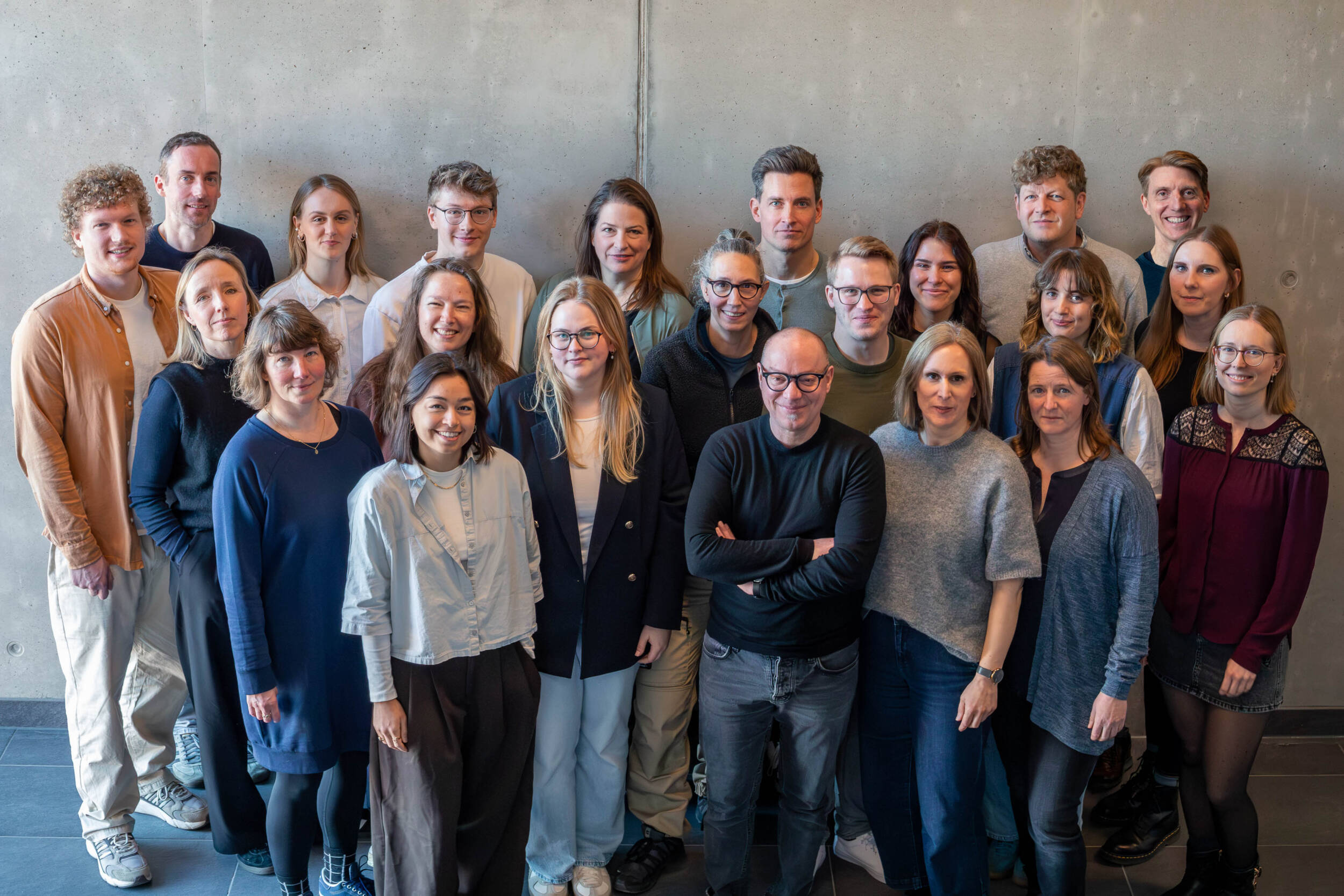 Gruppenporträt des MERA-Teams im Hamburger Büro für Landschaftsarchitektur. Die Mitarbeitenden stehen in mehreren Reihen vor einer ruhigen grauen Wand, blicken freundlich in die Kamera und sind natürlich als gemeinsames Team porträtiert.