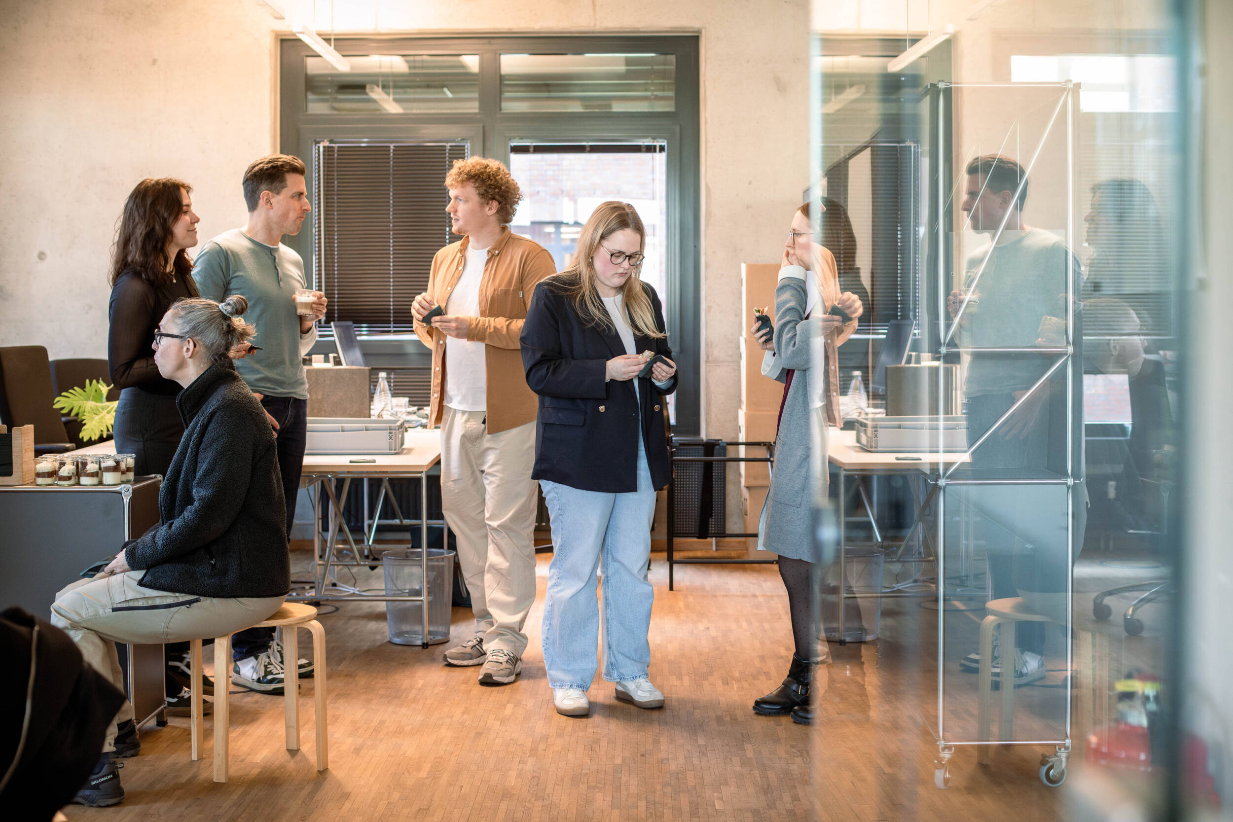 Mehrere MERA-Mitarbeitende kommen im offenen Hamburger Büro für Landschaftsarchitektur zu einem informellen Austausch zusammen. Einige stehen im Raum und unterhalten sich, eine Mitarbeiterin sitzt links an einem Tisch; im Hintergrund sind Arbeitsplätze, Glaswände und die helle Büroumgebung zu sehen.