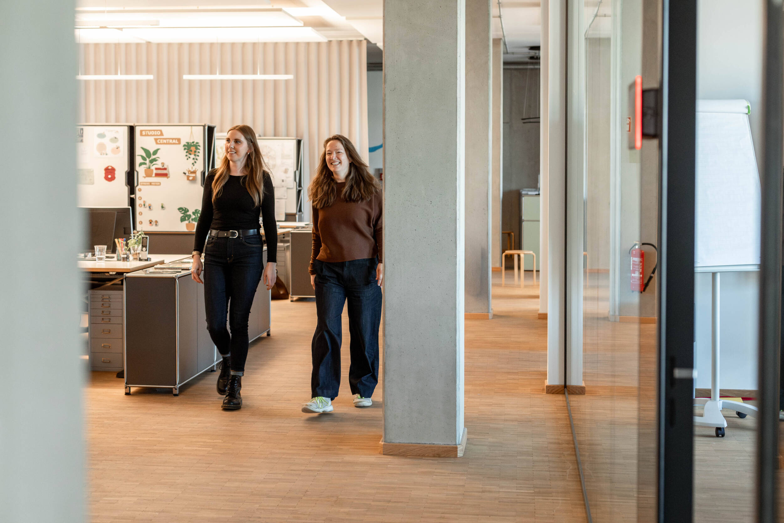 Zwei MERA-Mitarbeiterinnen stehen oder gehen gemeinsam durch das helle Hamburger Büro für Landschaftsarchitektur. Sie sind in einem offenen Arbeitsbereich mit Holzboden, Schreibtischen und Glaswänden zu sehen; im Hintergrund befinden sich Arbeitsplätze und Pinnflächen mit Notizen oder Plänen.