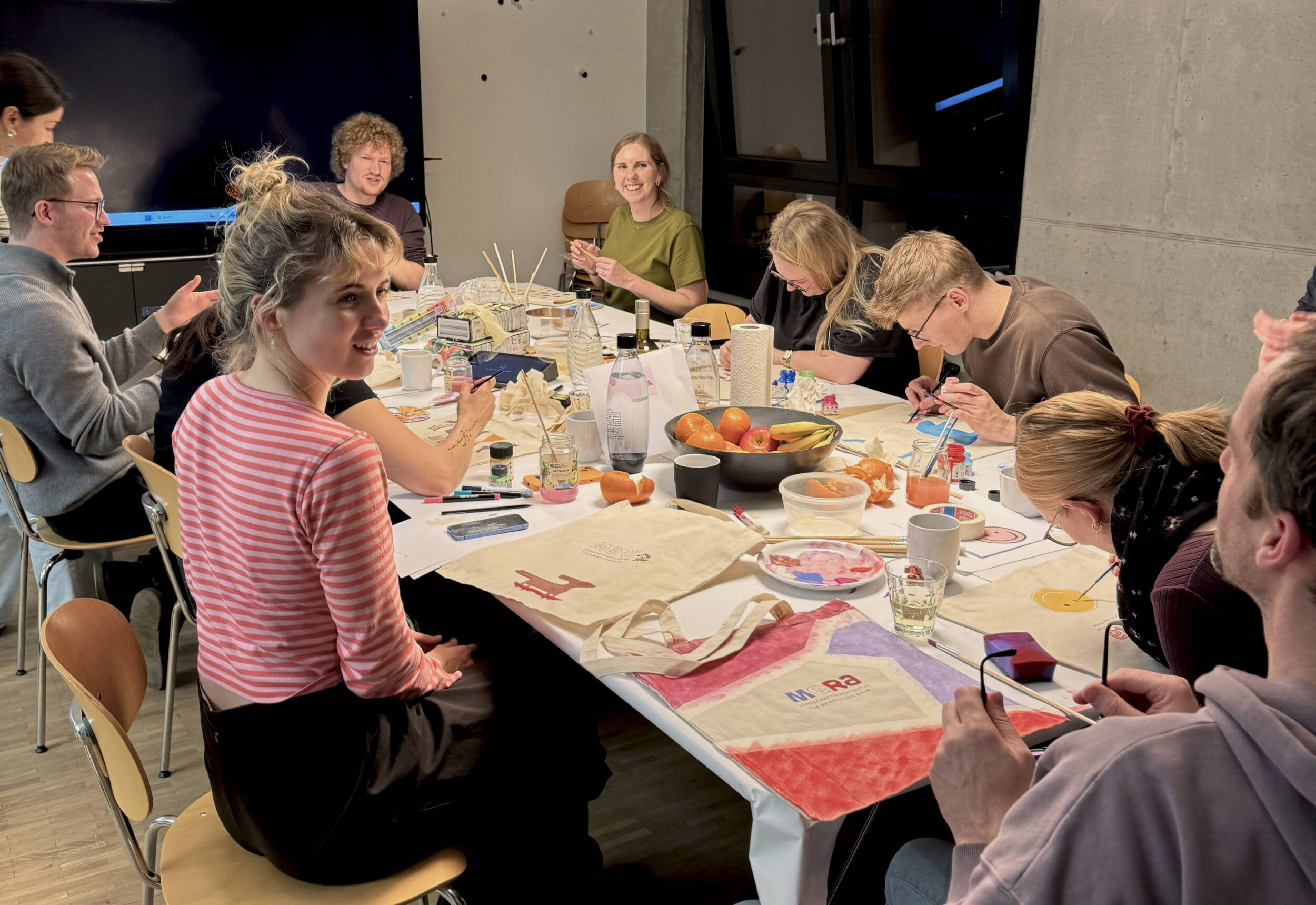 Das MERA-Team sitzt nach Feierabend im Hamburger Büro für Landschaftsarchitektur gemeinsam an einem großen Tisch und bemalt Stoffbeutel. Mehrere Mitarbeitende arbeiten mit Pinseln und Farben an eigenen Motiven, auf dem Tisch liegen bemalte Beutel, Farbteller, Stifte, Gläser und Obst. Die Szene wirkt kreativ, ungezwungen und gemeinschaftlich.