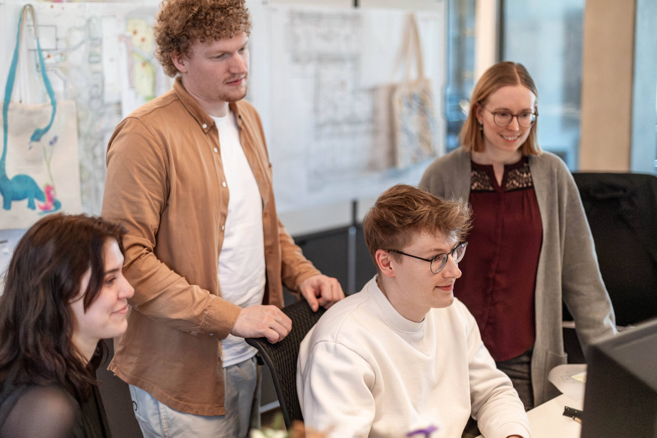 Vier Kolleg*innen von MERA arbeiten gemeinsam an einem Bildschirm; im Hintergrund hängen Planzeichnungen