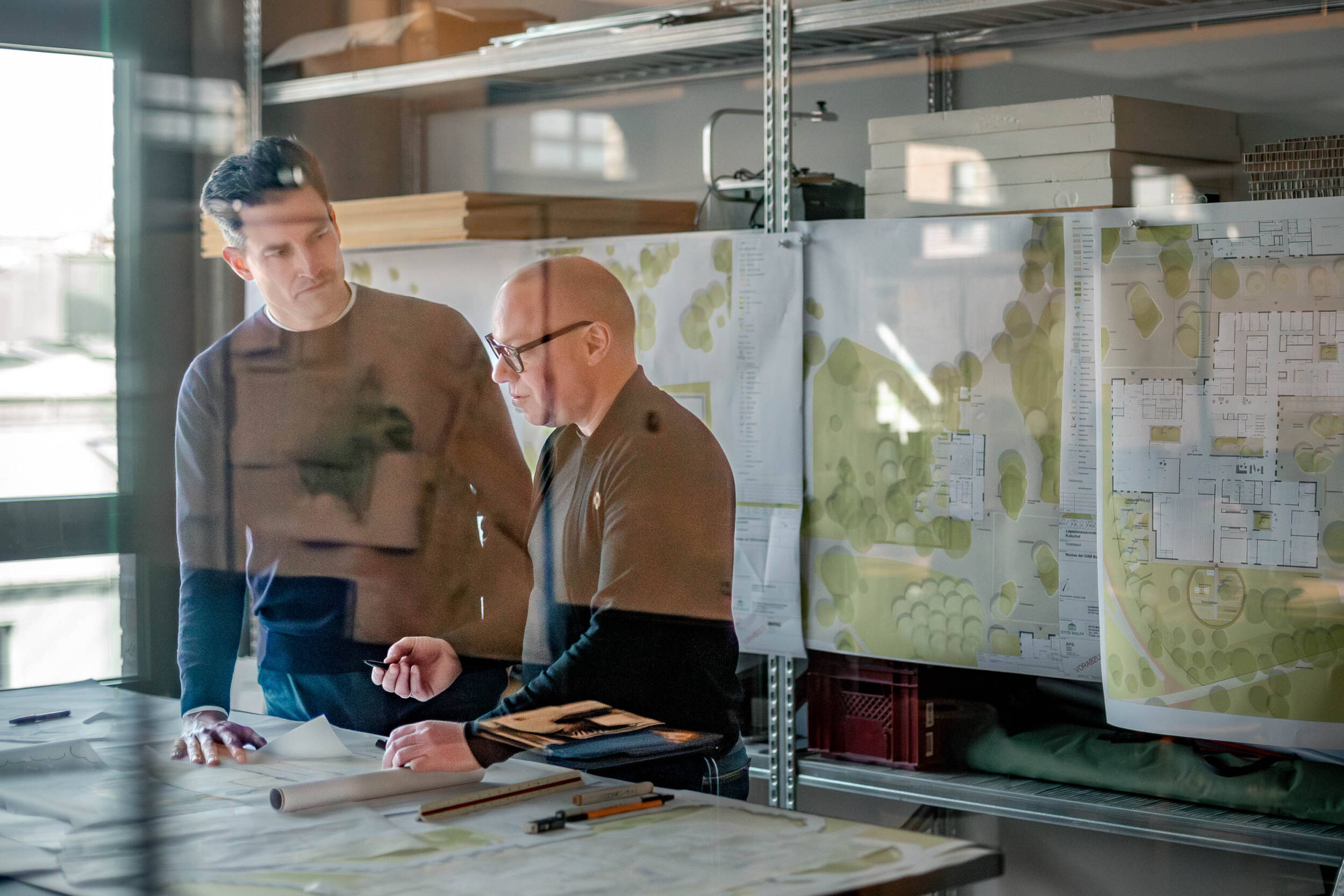 Arbeitssituation im MERA-Büro: Zwei Personen besprechen Planunterlagen an einem großen Arbeitstisch; im Hintergrund hängen und liegen großformatige Freiraumpläne an einem Regal, die Aufnahme ist durch eine Glasscheibe fotografiert.