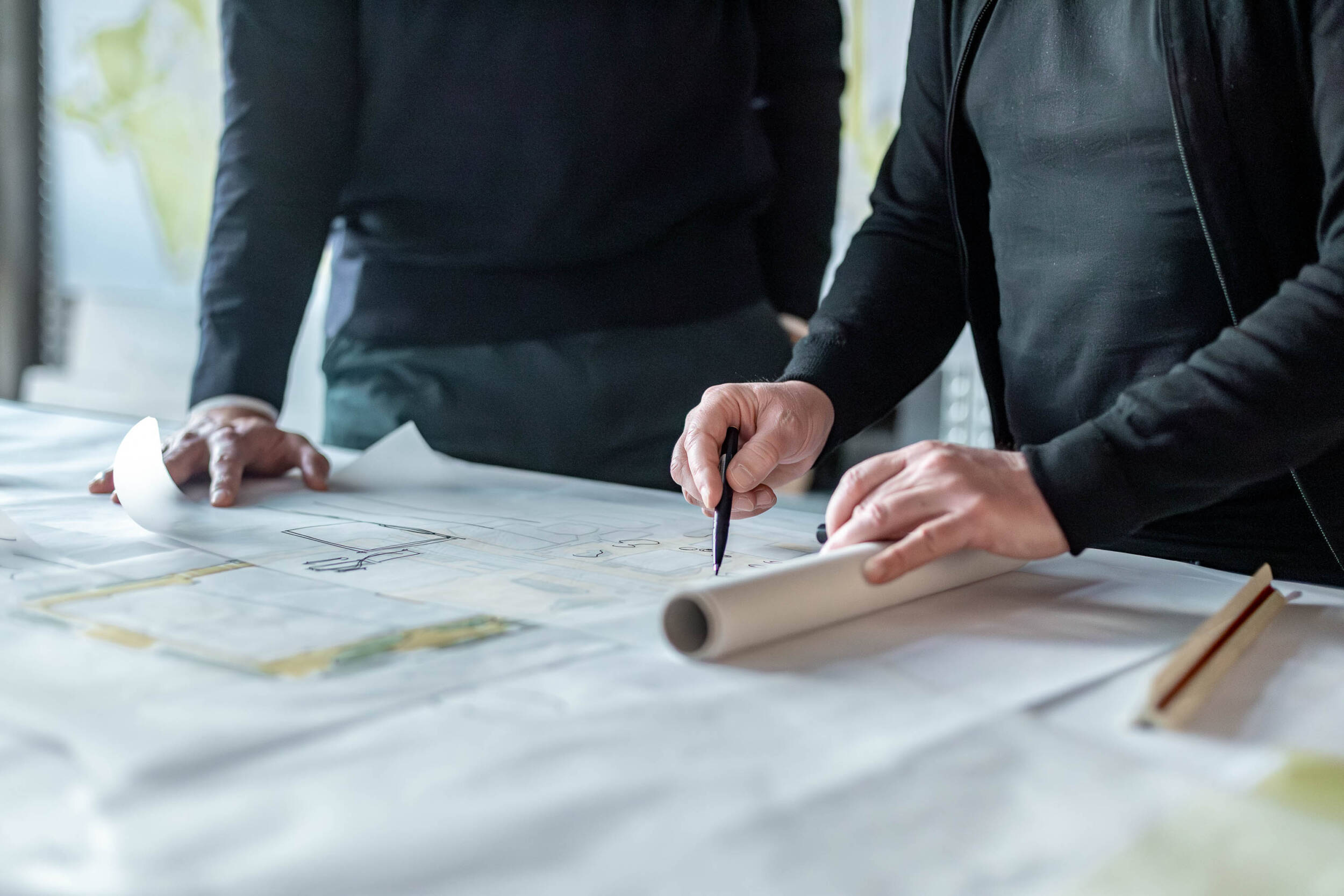Planungssituation bei MERA: Zwei Personen arbeiten an großformatigen Planunterlagen, eine Hand markiert Details mit einem Stift, daneben liegen gerollte Pläne und Zeichenwerkzeuge.