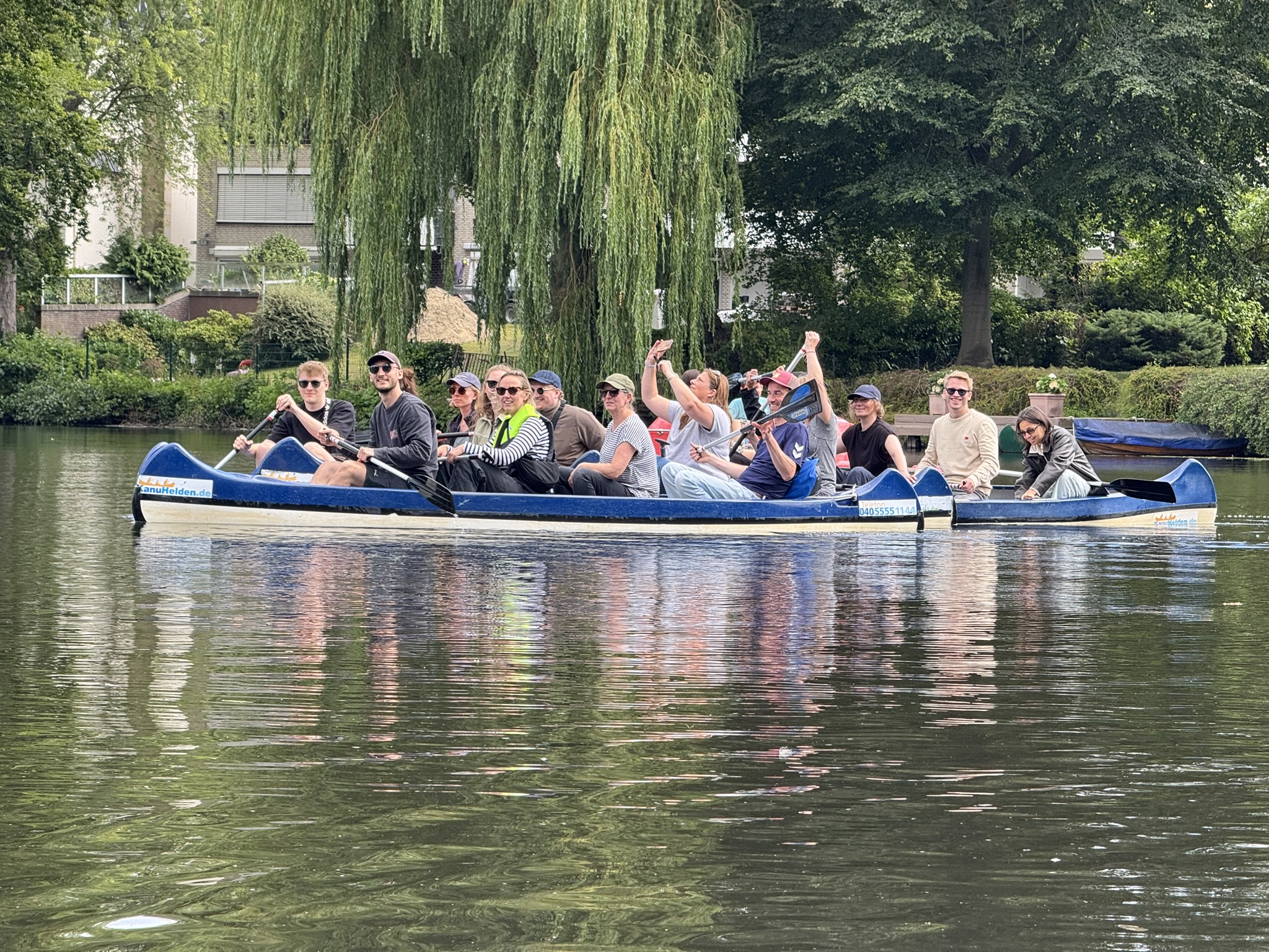 Zu sehen ist das gesamte Team MERA bei einem fröhlichen Kanuausflug auf der Hamburger Alster. Die Stimmung scheint ausgelassen, sommerlich, alle tragen Sonnenbrillen. im Hintergrund ist üppiger Baumbestand zu sehen, im Bild Vordergrund sind die Spiegelungen der Kanus und der Personen auf dem Wasser zu sehen.
