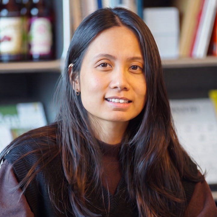 Porträt von Yi Li aus dem MERA-Team. Sie blickt mit einem leichten Lächeln direkt in die Kamera. Ihre langen, dunklen Haare trägt sie offen, dazu ein dunkelbraunes Oberteil. Im unscharfen Hintergrund sind Bücherregale der Büroumgebung zu erkennen.